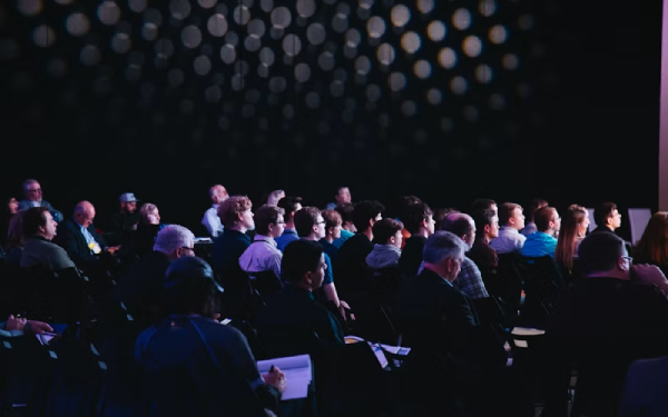 An audience seated in a dimly lit hall watching a live presentation.